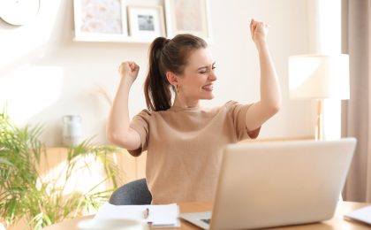 Happy entrepreneur woman sit at desk reading good news and express joy rising hands up.