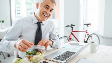 Happy businessman opening his salad pack and having a lunch break at office desk enjoying healthy eating